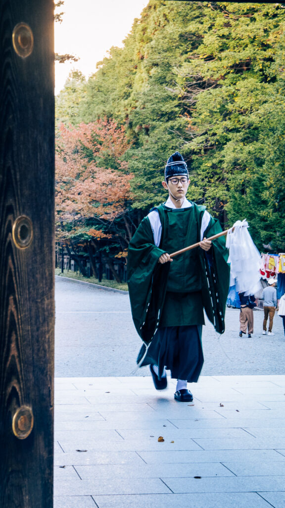 A Shinto clergy member in green robes walks into the shrine area carrying a harai gushi purification wand at Hokkaido-Jingu