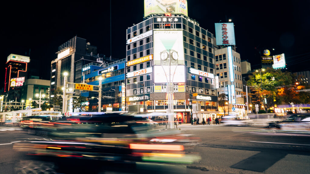 Night traffic streaks across a large Sapporo intersection in long exposure beneath tall neon signs.