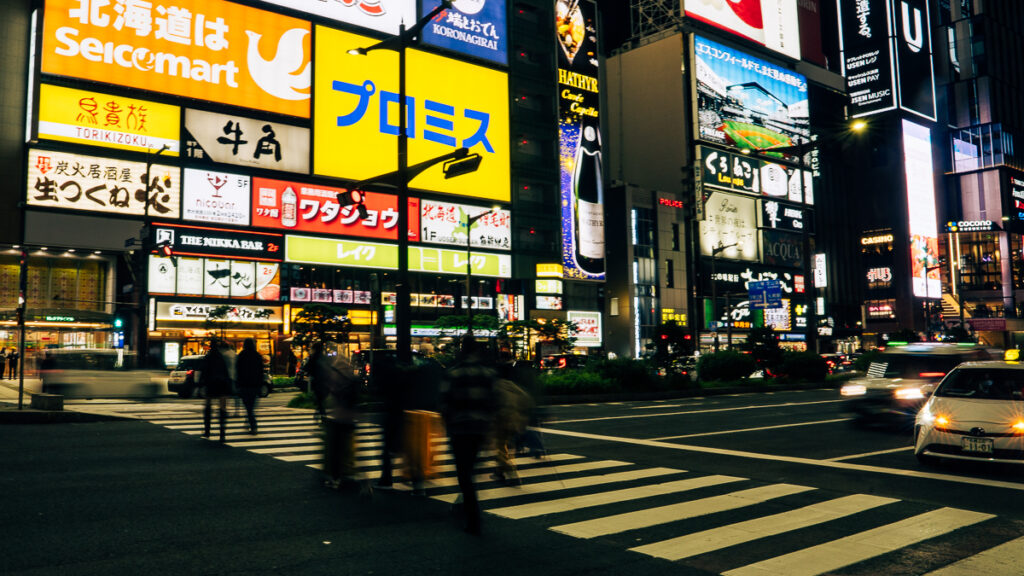 Long-exposure pedestrians walking across a busy Sapporo intersection under bright neon signage, Hokkaido.
