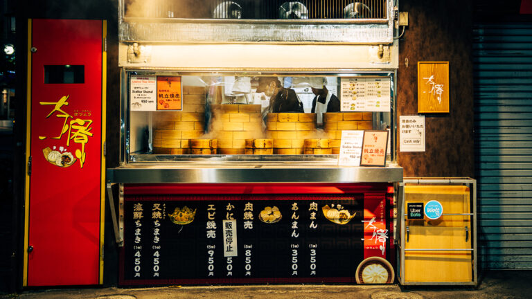 Steam rising from bamboo steamer baskets at a red street food stall in Sapporo at night, workers visible through a small window.