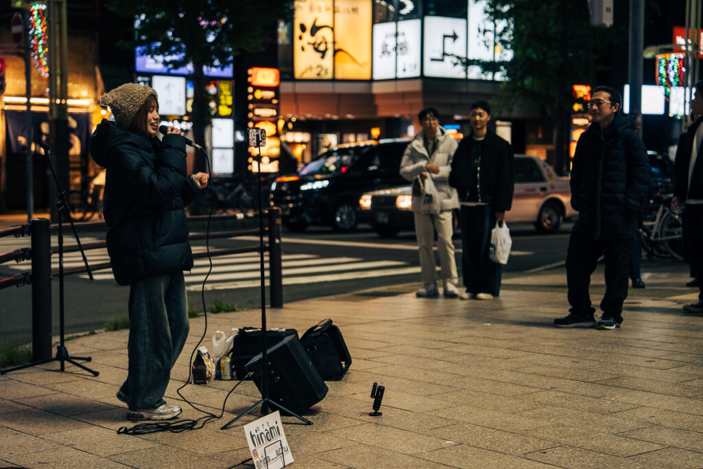 Street musician singing on a corner in Sapporo with a mic, speaker, and camera set to record, small audience watching.