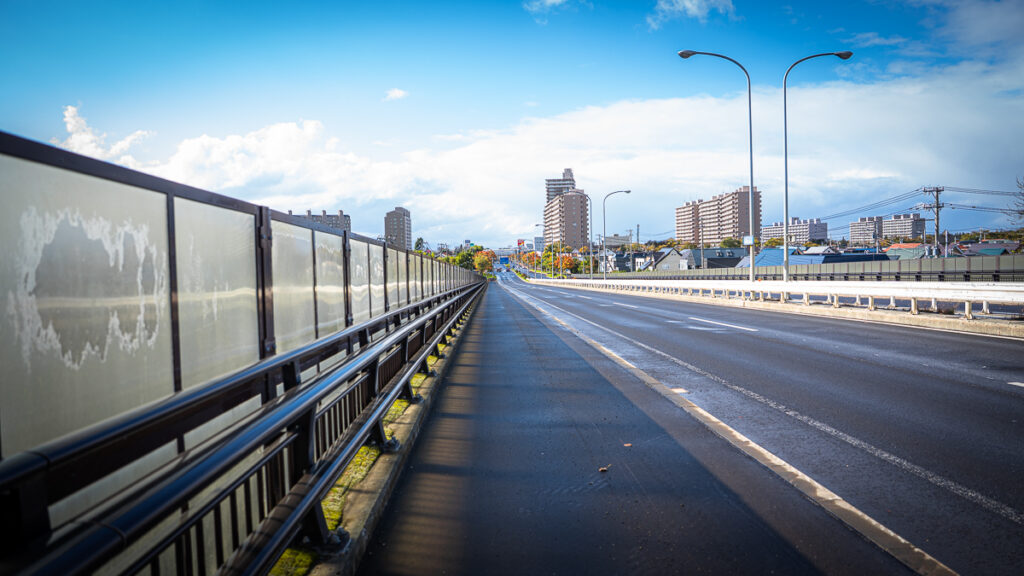 A wide, empty suburban road in Sapporo with tall apartments visible in the distance.