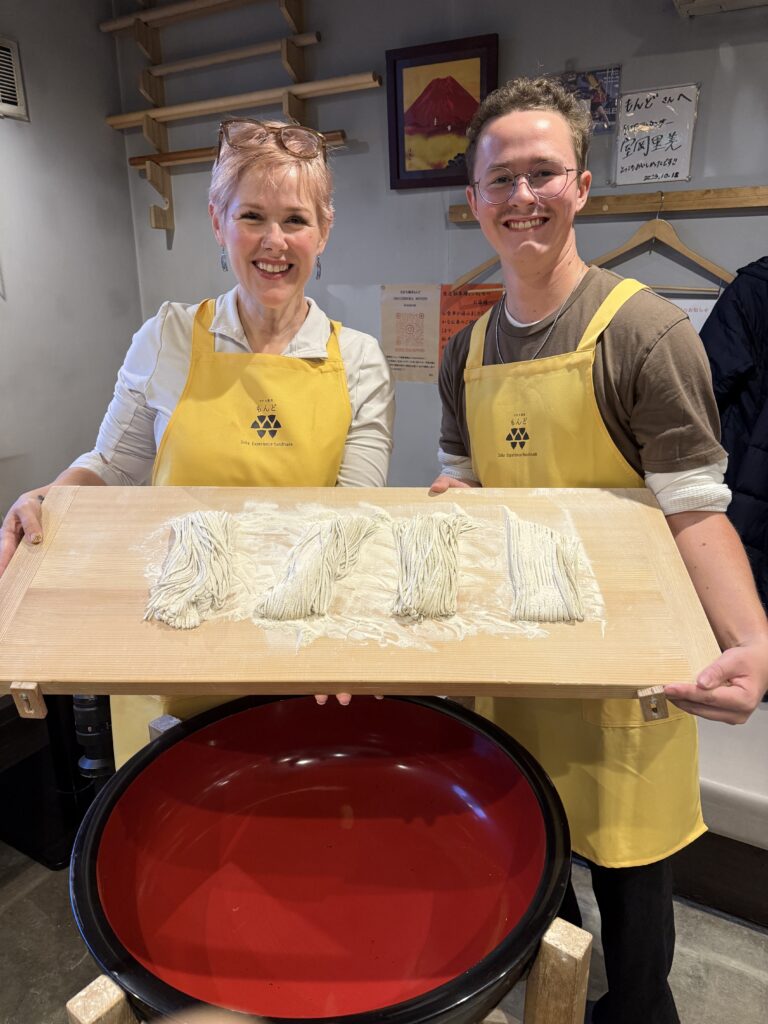 Mac and Laurie smiling and holding up a board of freshly cut soba noodles while wearing yellow aprons.
