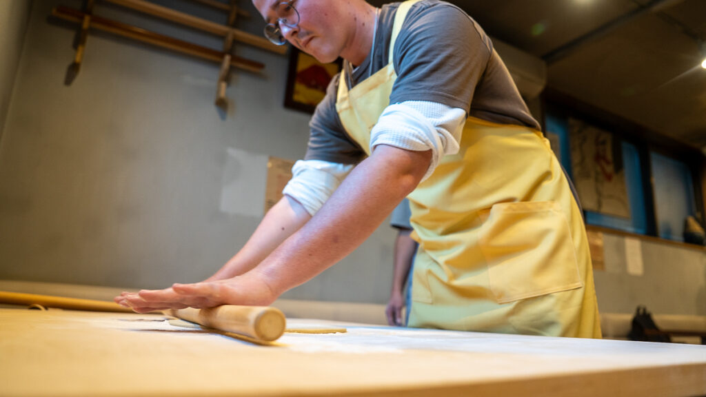 Mac uses a long roller to gently stretch soba dough on a wooden worktable.