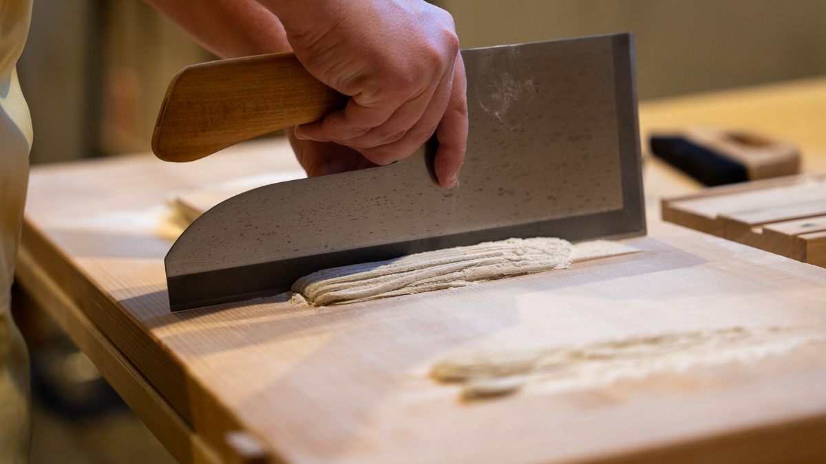 Close-up of a soba knife slicing thin buckwheat noodles into precise strips.