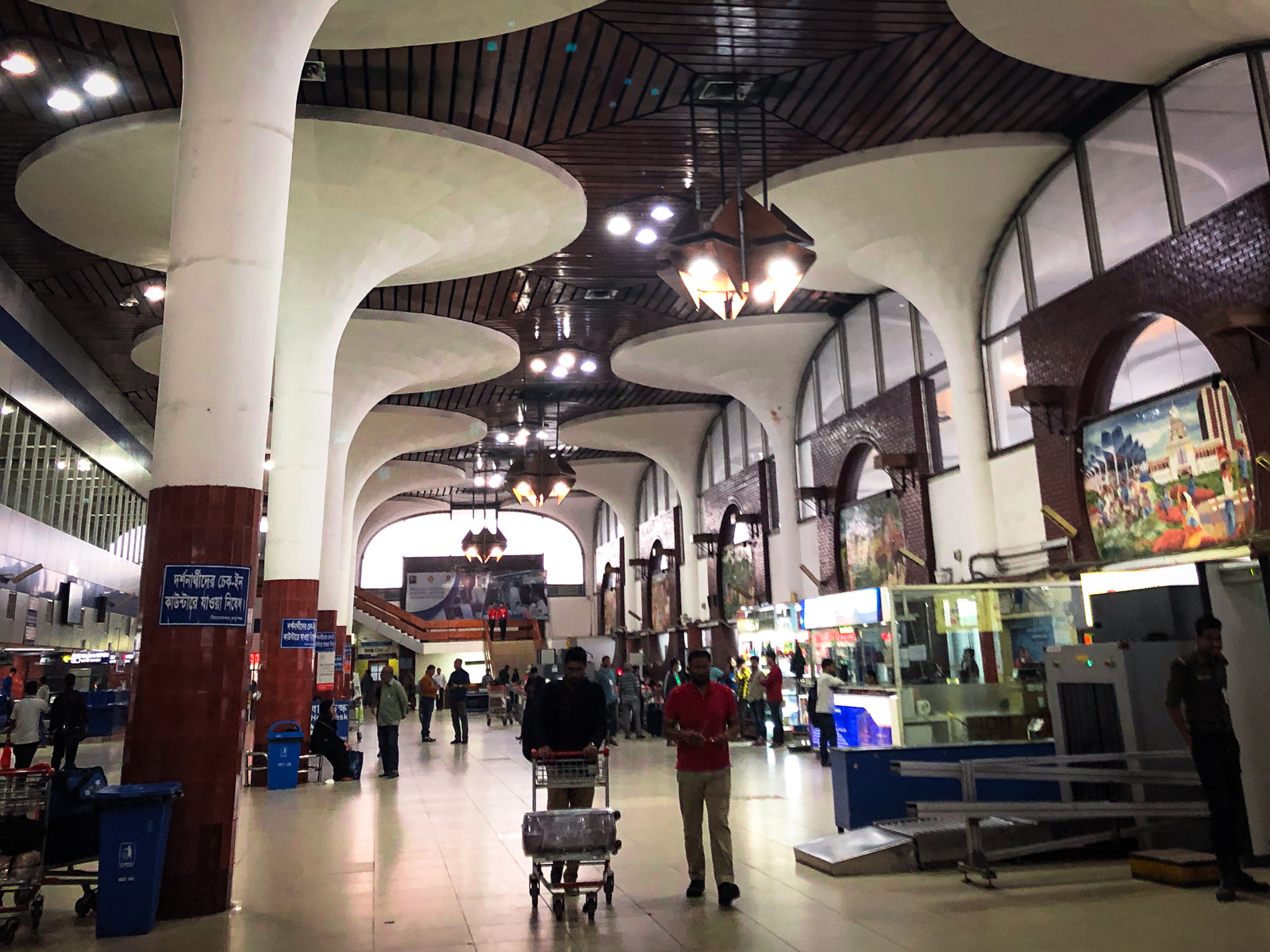 Interior shot of Dhaka airport, large pillars reach to the ceiling and two people move with a luggage cart down the aisle