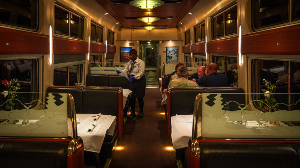 Wide evening view inside an Amtrak dining car, with four passengers seated at one table and a train employee clearing another table as the train travels across the country.