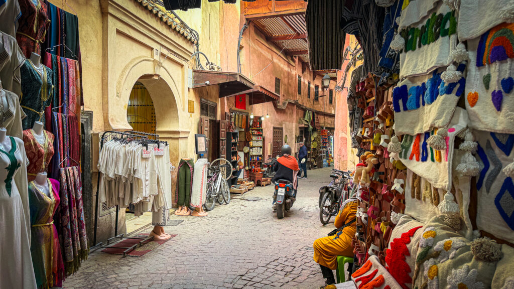 Motorcycle navigating a narrow Marrakech medina street past vendors selling rugs and textiles