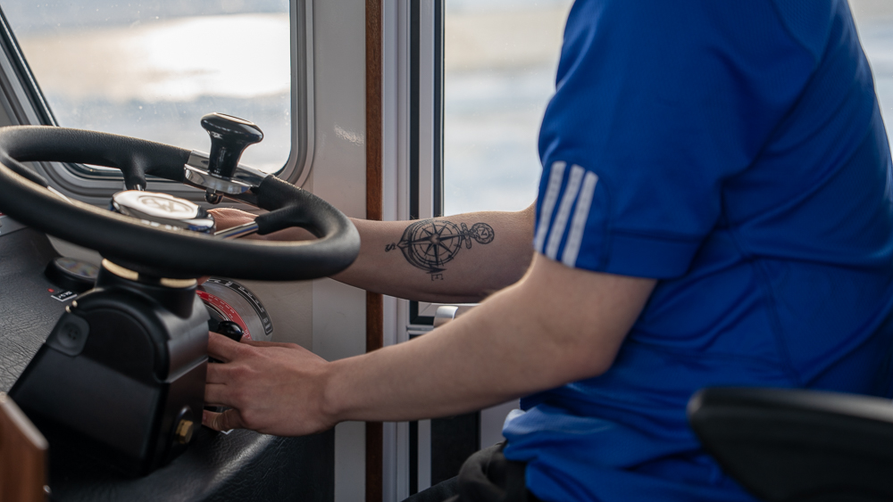 Close-up of a boat captain steering an iceberg tour vessel, compass tattoo visible on his arm.