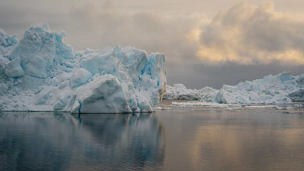 Blue and white iceberg reflected in calm water beneath sunset-lit clouds in Greenland.