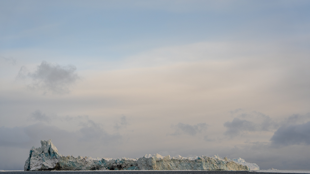 Extremely long iceberg with peaks, brown sediment streaks, and blue ice in Disko Bay.