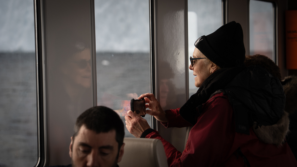 Passengers looking through windows of an iceberg tour boat with reflections visible on metal walls.