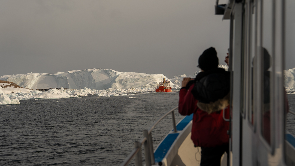 Tourist leaning from a boat doorway to photograph a red fishing boat passing an iceberg in Disko Bay.