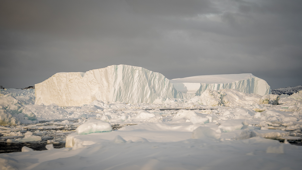 White iceberg floating beside flat sea ice beneath gray Arctic skies.