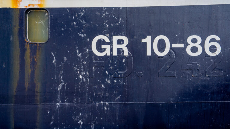Close-up of a navy blue ship hull marked "GR 10-86," with rust streaks and white splatters across the painted surface.