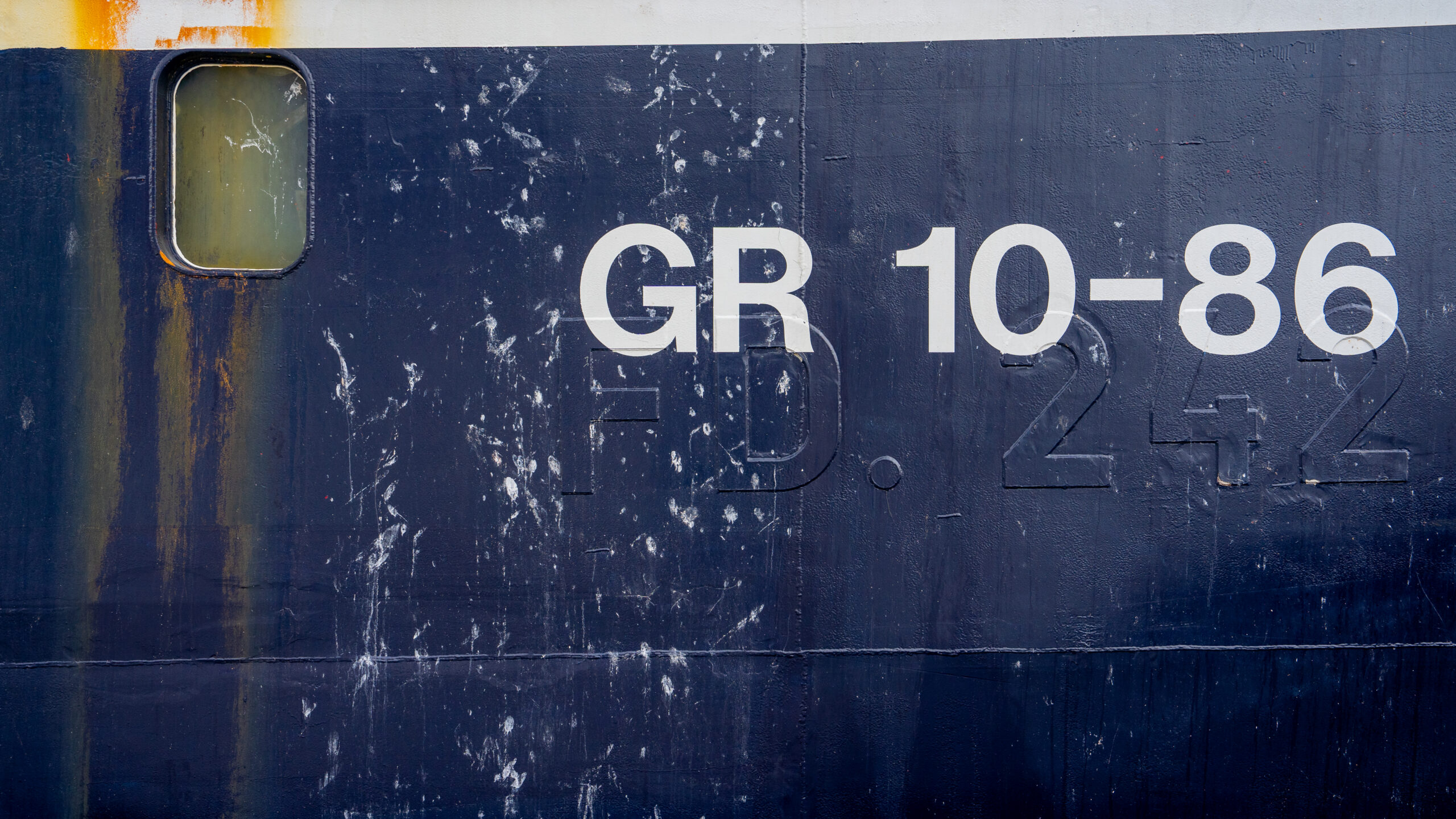 Close-up of a navy blue ship hull marked "GR 10-86," with rust streaks and white splatters across the painted surface.
