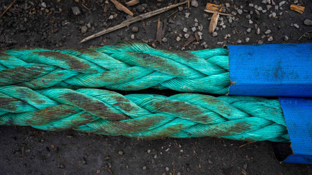 Close-up of thick turquoise braided rope resting against dark brown ground with blue protective casing along one side.