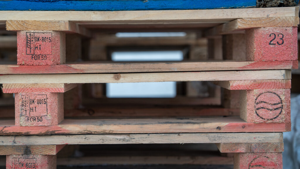 Close-up of stacked wooden pallets with faded reddish ends marked with identification numbers and logos.