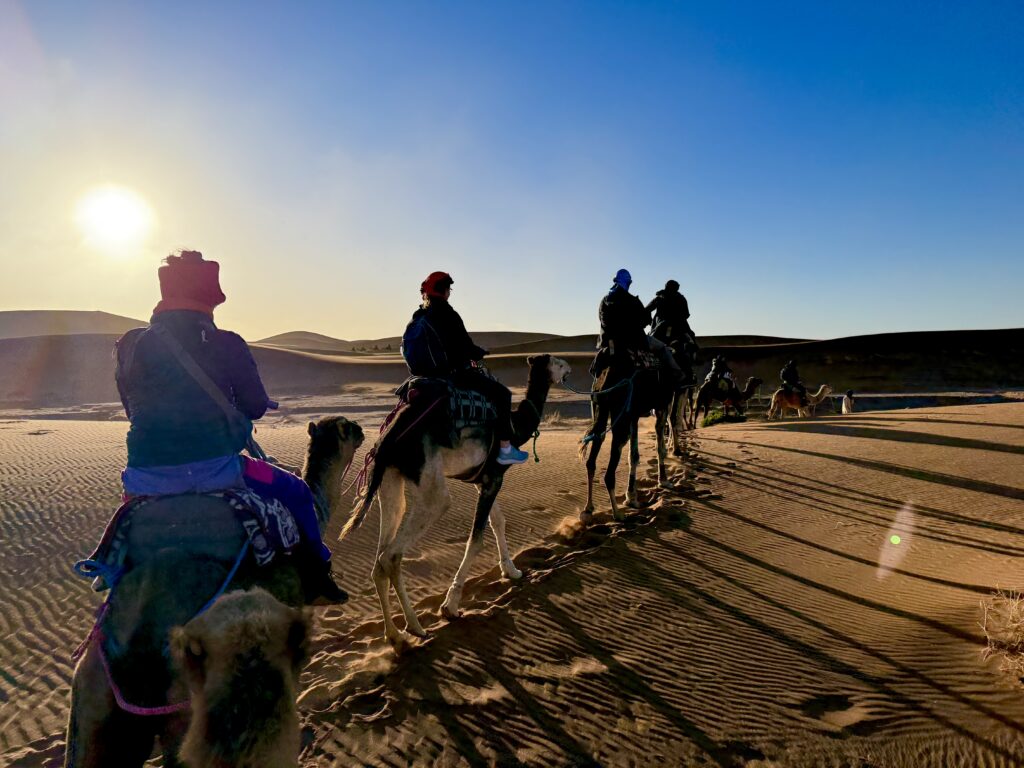 A line of camel riders crosses terracotta-colored dunes under a clear blue sky near Erg Chigaga in Morocco.