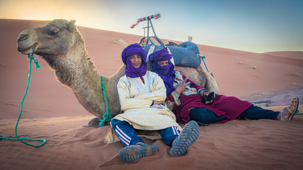 Two camel keepers lean against a seated camel while waiting near the dunes of Erg Chigaga in the Sahara Desert.