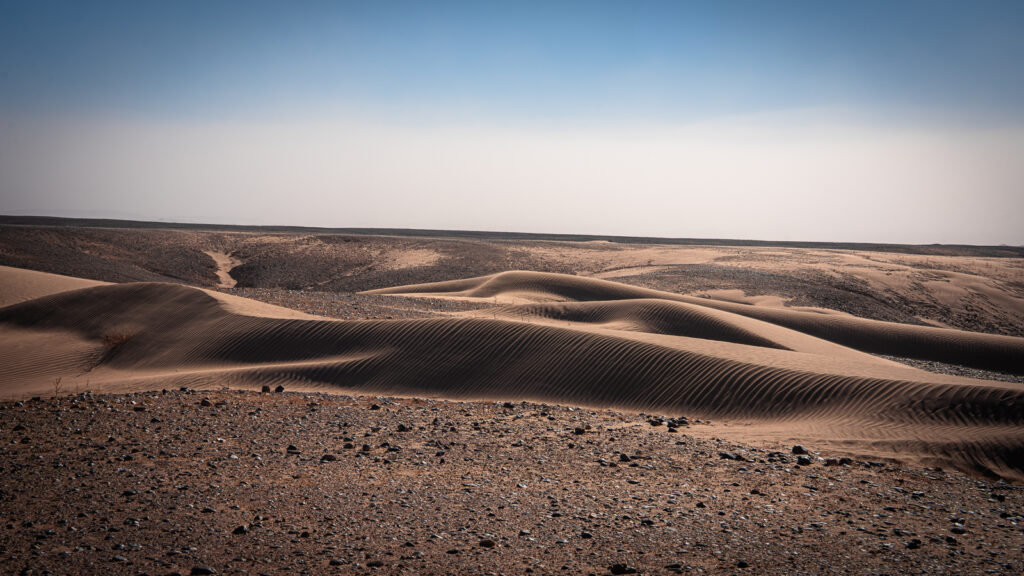 Wide view of desert landscape near Erg Chigaga showing different textures of sand and rocky gravel across the surface.
