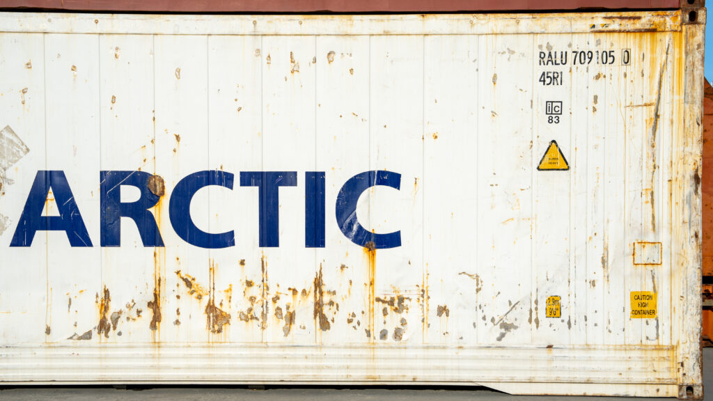 Close-up of a white shipping container with large blue letters spelling "Arctic" and chipped paint exposing rust beneath.