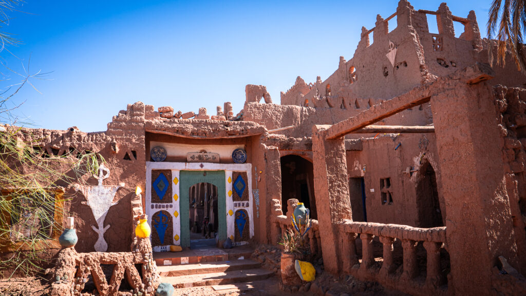 Entrance to Auberge Kasbah Touareg in M’Hamid, Morocco, with red clay walls, intricate roof detailing, a green door painted with white and blue patterns, and yellow pots beside the entry.