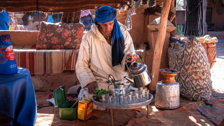Older man in white djellaba and blue head wrap prepares Moroccan tea while seated on the floor beside a tray of small glasses, silver teapot, and tins of herbs and sugar at Auberge Kasbah Touareg in M’Hamid.