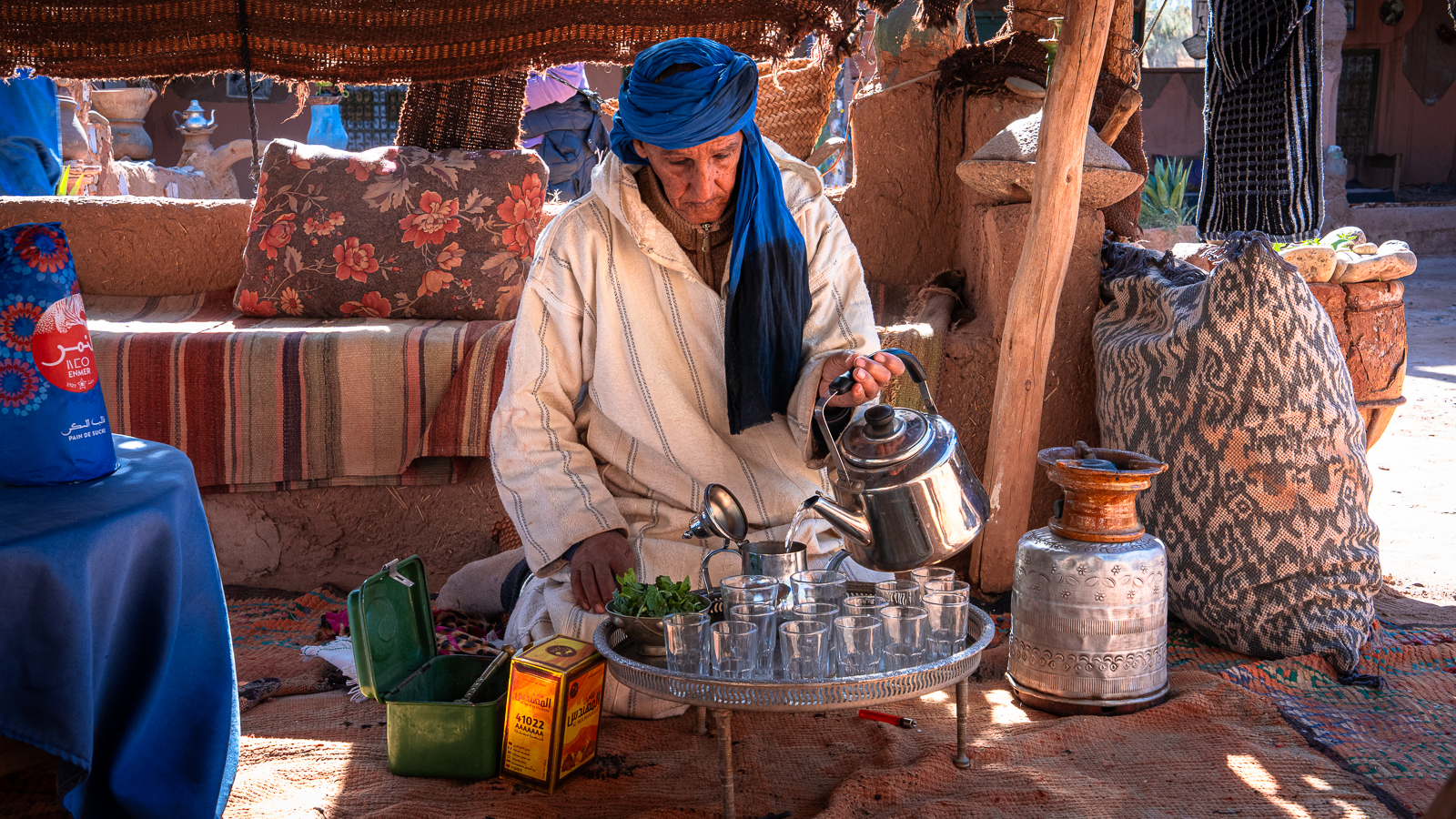 Older man in white djellaba and blue head wrap prepares Moroccan tea while seated on the floor beside a tray of small glasses, silver teapot, and tins of herbs and sugar at Auberge Kasbah Touareg in M’Hamid.