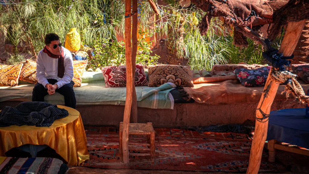 Traveler resting on cushioned benches in the shade inside the tented seating area at Auberge Kasbah Touareg in M’Hamid, Morocco.