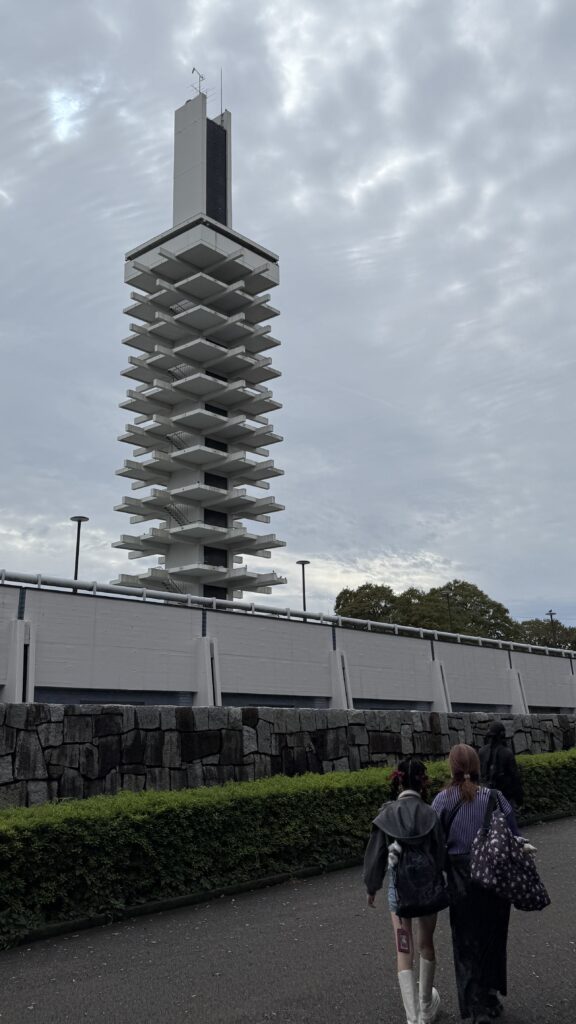 Two women walk toward Tokyo Ramen Festa with Komazawa Olympic Park Tower in the background.