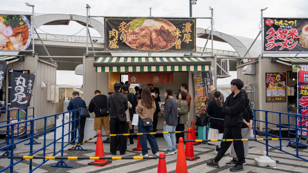 Festival visitors stand in line at the Saga Miso Noodle Club booth serving rich miso ramen topped with flame-grilled pork belly.