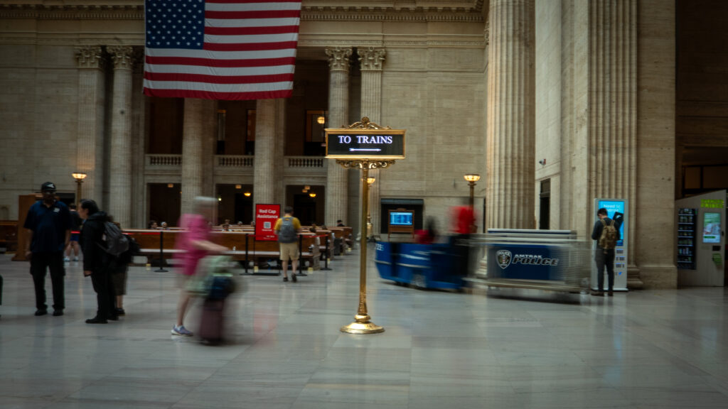 Blurred long-exposure of passengers rushing by a “To Trains” directional sign at Chicago Union Station.