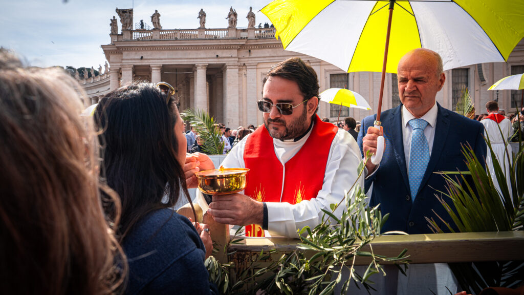 Priest placing host into mouth of worshipper while assistant holds umbrella in Vatican