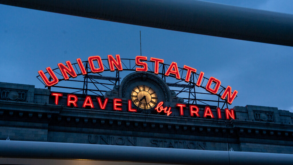 Neon Union Station sign in Denver with its analog clock and the words “Travel by Train.”
