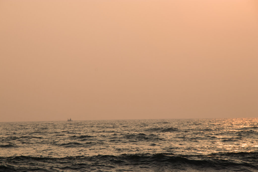 The faint silhouette of a moon boat appears in the distance beneath a pink sunset over the Bay of Bengal.