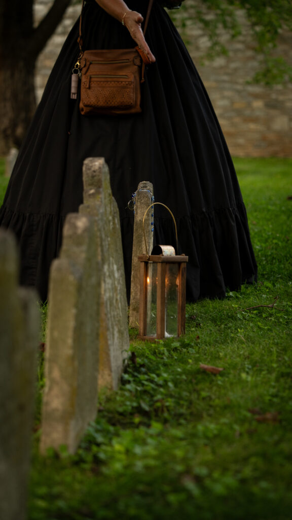 Old cemetery headstones and a lantern stand beside the black skirt of a Shepherdstown Mystery Walk guide.