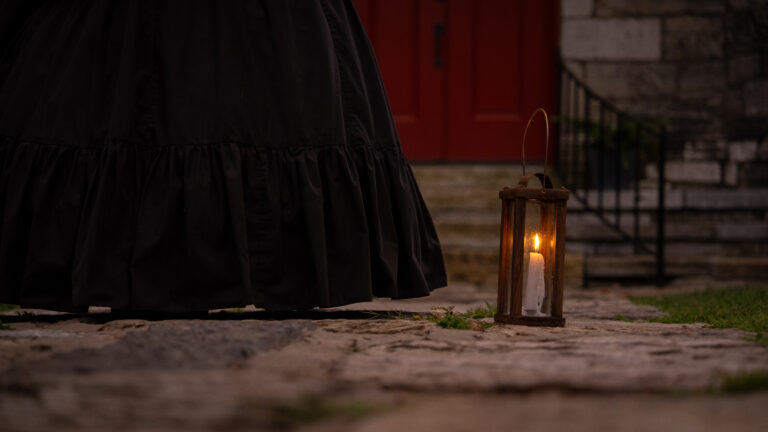 A glowing lantern rests on a cobblestone walk beside a guide in a black skirt, with an old stone church and red door behind her.