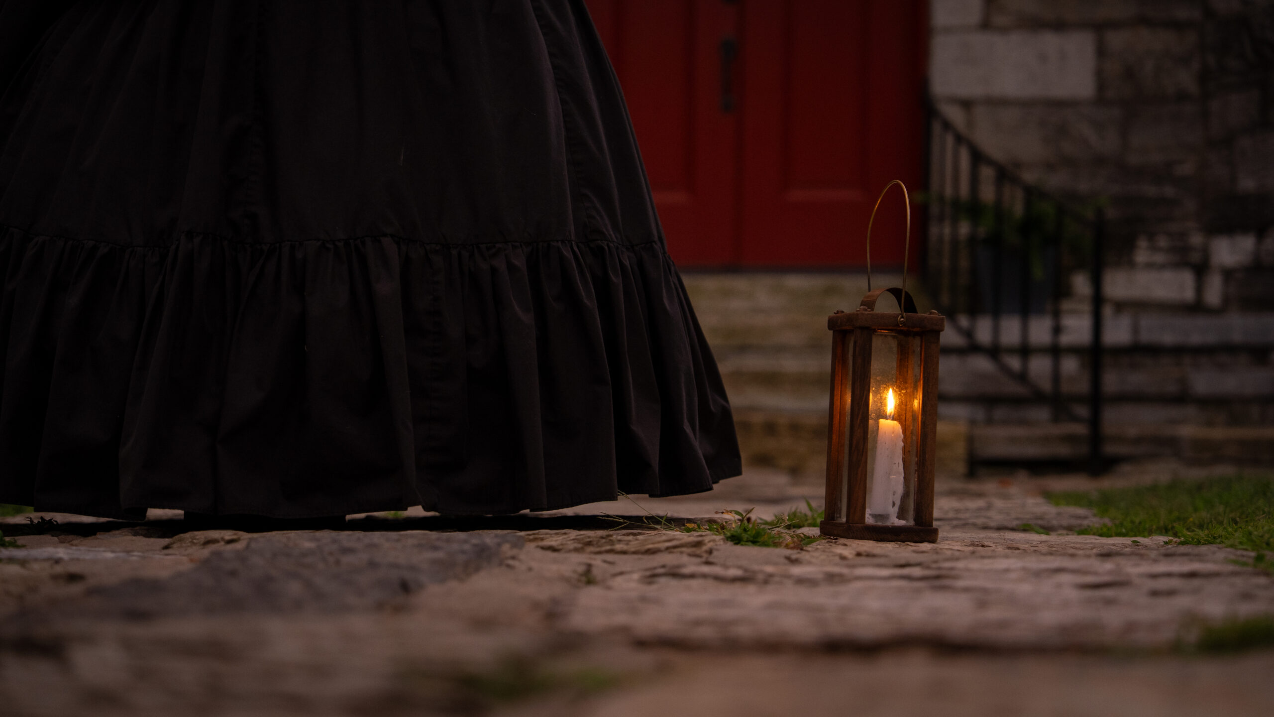 A glowing lantern rests on a cobblestone walk beside a guide in a black skirt, with an old stone church and red door behind her.