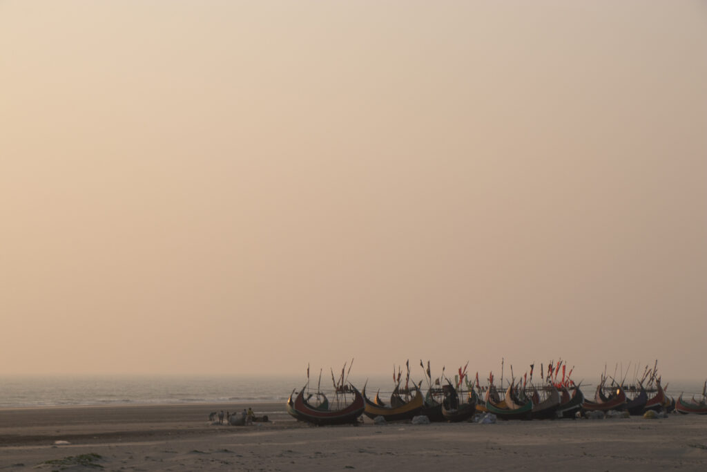 About a dozen moon boats line a wide beach beneath an orange-pink sunset on Bangladesh’s southeastern coast.