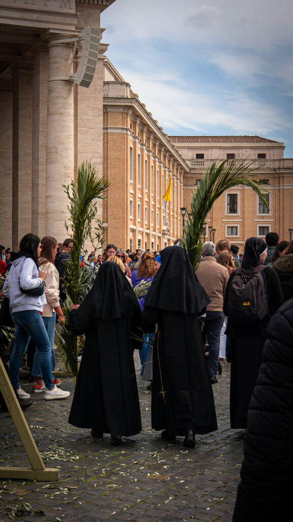 Two nuns in black habits walking away carrying large palm branches after Mass