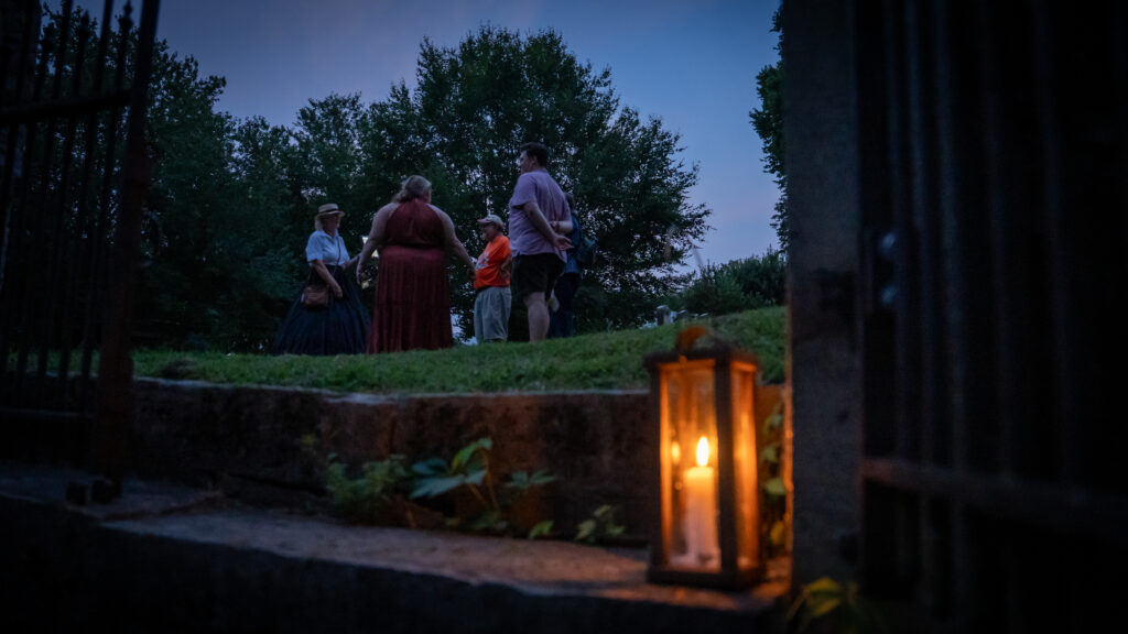A glowing lantern sits on a stone step while Shepherdstown Mystery Walk guests gather in a circle in the background.