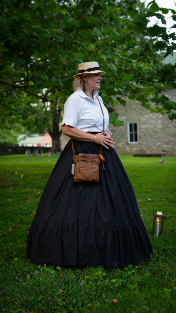 Janet Hughes in a black hoop skirt stands in a historic Shepherdstown cemetery with a stone church behind her.