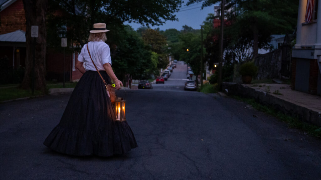 A lantern carried by a walking-tour guide glows on a dark Shepherdstown street at dusk.