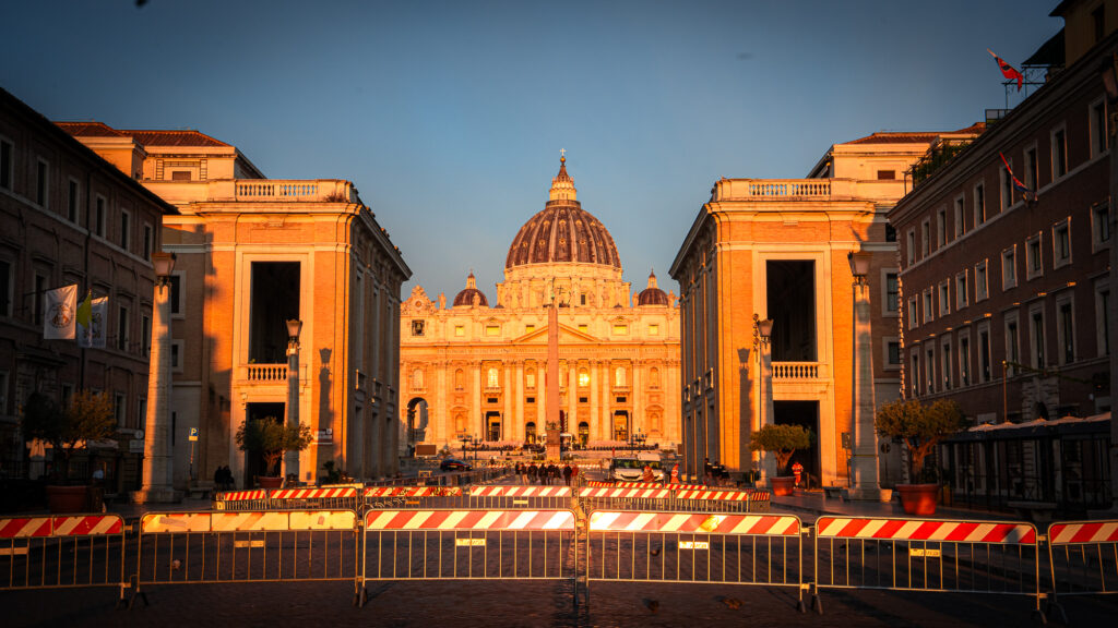 Sunrise illuminating St Peter’s Basilica behind gates and lines of people entering the square